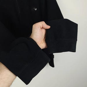 Puede incluir: Camisa negra de manga larga con cuello alto y un peque&ntilde;o detalle de bot&oacute;n. Los pu&ntilde;os son gruesos y doblados. La camisa est&aacute; hecha de una tela suave y texturizada.