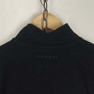 Puede incluir: Jersey polar negro con cuello alto. La palabra "MAMMUT" est&aacute; bordada con hilo oscuro en el pecho. La prenda est&aacute; colgada de una percha de madera con una cadena de metal.