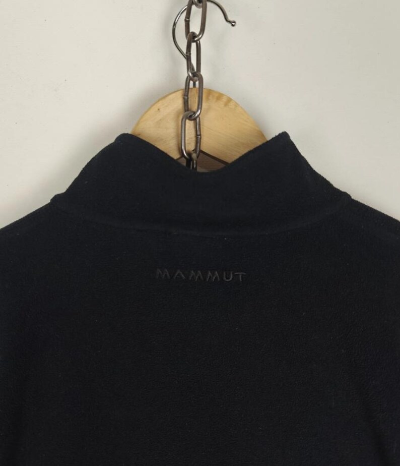 Puede incluir: Jersey polar negro con cuello alto. La palabra "MAMMUT" est&aacute; bordada con hilo oscuro en el pecho. La prenda est&aacute; colgada de una percha de madera con una cadena de metal.