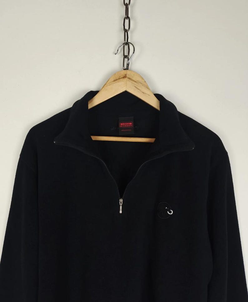 Puede incluir: Sudadera negra de manga larga con cuello con cremallera de un cuarto. La sudadera tiene un peque&ntilde;o logotipo en el pecho y est&aacute; colgada de una percha de madera. El fondo es de color blanquecino.