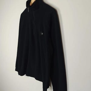 Puede incluir: Jersey negro de manga larga con cuello alto y cierre de cremallera de un cuarto. La camisa tiene un peque&ntilde;o logotipo circular en el pecho. La prenda est&aacute; colgada de una percha de madera suspendida de una cadena de metal. El fondo es una pared lisa blanquecina.