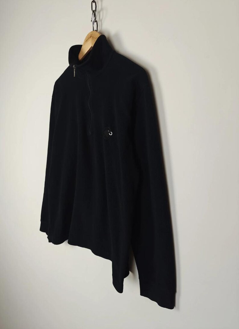 Puede incluir: Jersey negro de manga larga con cuello alto y cierre de cremallera de un cuarto. La camisa tiene un peque&ntilde;o logotipo circular en el pecho. La prenda est&aacute; colgada de una percha de madera suspendida de una cadena de metal. El fondo es una pared lisa blanquecina.