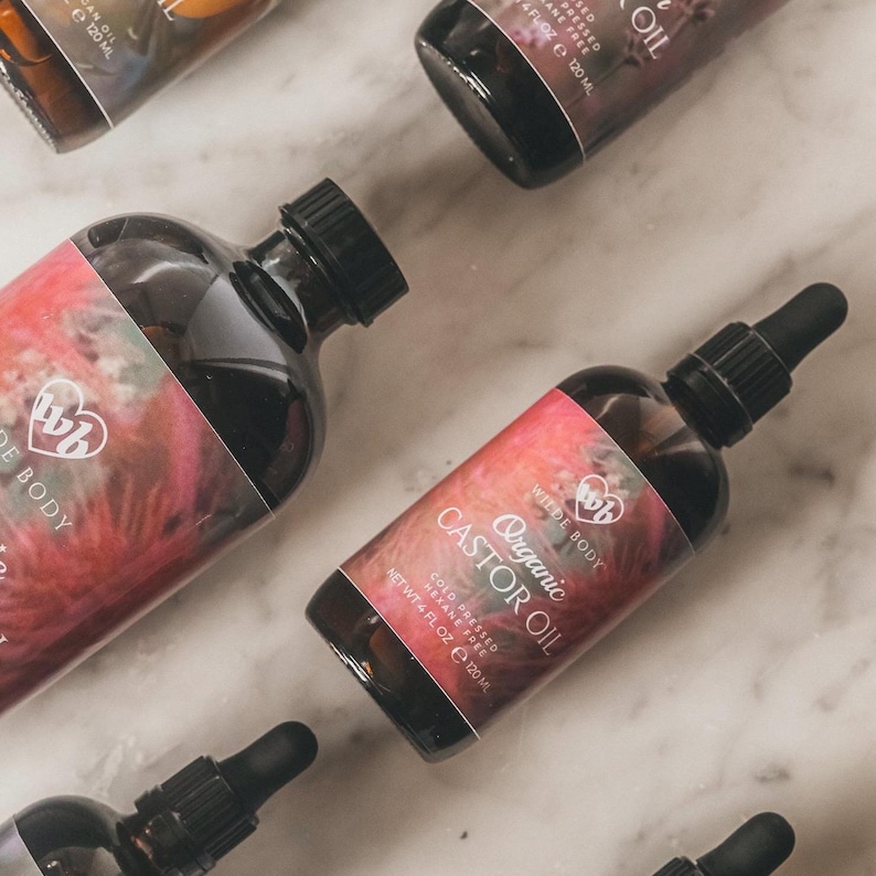 May include: Three glass bottles of organic castor oil, cold pressed and hexane free. The bottles are labeled with the brand name "Wilde Body" and the words "Organic Castor Oil". The bottles are on a white marble surface.