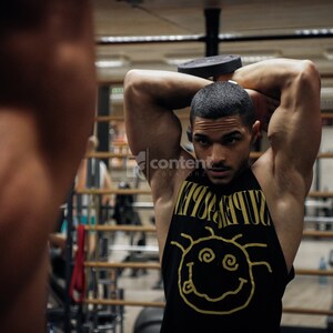 May include: A man in a black tank top with a yellow graphic of a smiley face and the word "SUPREME" is lifting a black dumbbell over his head in a gym.