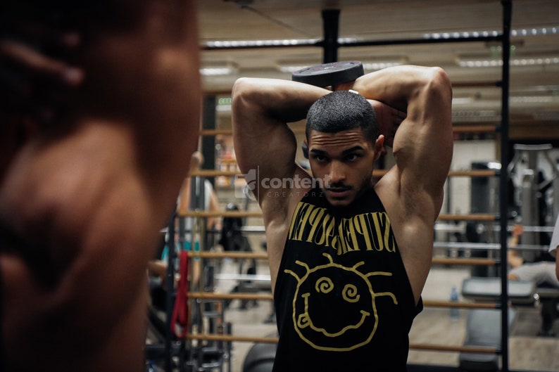May include: A man in a black tank top with a yellow graphic of a smiley face and the word "SUPREME" is lifting a black dumbbell over his head in a gym.