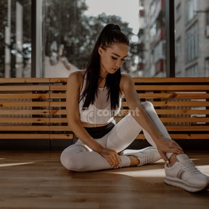 May include: A woman wearing a white tank top and white leggings sits on a wooden bench with her legs crossed. She is wearing white sneakers and has her hair pulled back in a ponytail.