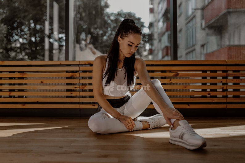 May include: A woman wearing a white tank top and white leggings sits on a wooden bench with her legs crossed. She is wearing white sneakers and has her hair pulled back in a ponytail.
