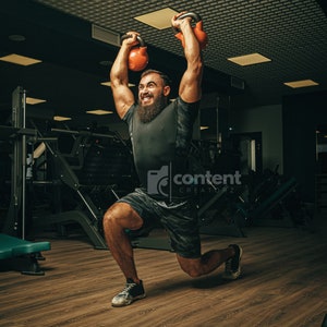 May include: A bearded man wearing a black shirt and camouflage shorts performs a lunge exercise while holding two orange kettlebells overhead in a gym.