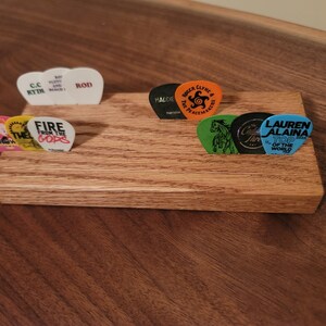 May include: A wooden guitar pick holder with eight guitar picks. The picks have various designs and text, including "Fire from the Gods", "C.C Rydi", "Rod", "Lauren Alaina Top of the World", "The Peacemakers", "Calder Thre", and "Crow".