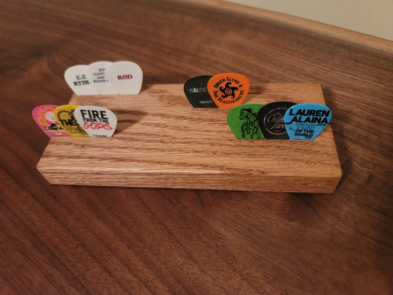 May include: A wooden guitar pick holder with eight guitar picks. The picks have various designs and text, including "Fire from the Gods", "C.C Rydi", "Rod", "Lauren Alaina Top of the World", "The Peacemakers", "Calder Thre", and "Crow".