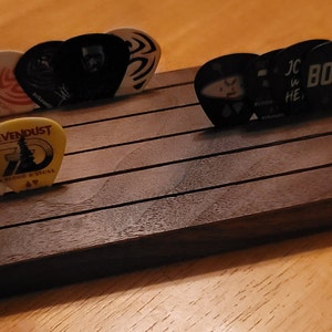 May include: A wooden guitar pick holder with a dark brown finish. The holder has five slots for storing guitar picks. There are several guitar picks in the holder, including picks with the words "SevenDust", "Boss", and "JC HE" on them.