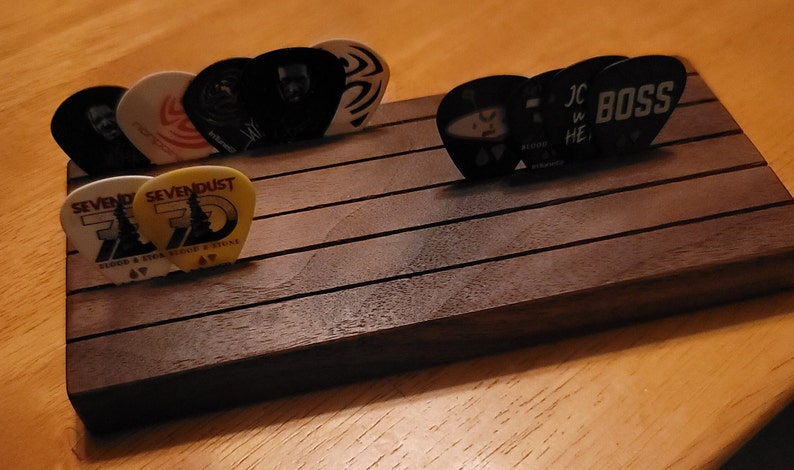 May include: A wooden guitar pick holder with a dark brown finish. The holder has five slots for storing guitar picks. There are several guitar picks in the holder, including picks with the words "SevenDust", "Boss", and "JC HE" on them.
