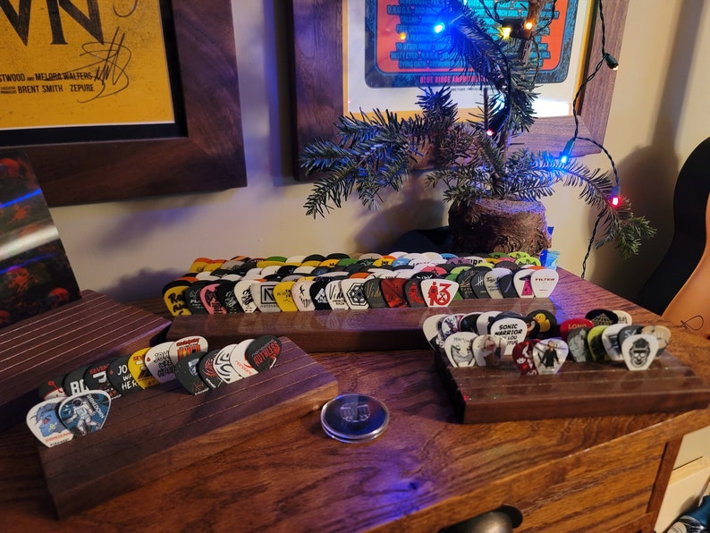 May include: Two wooden guitar pick holders with a variety of colorful guitar picks. The holders are on a wooden surface.