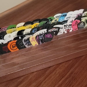 May include: A wooden guitar pick holder with a clear plastic front, displaying a collection of colorful guitar picks with various band logos and names. The picks are arranged in a fan shape, with the holder sitting on a wooden surface.