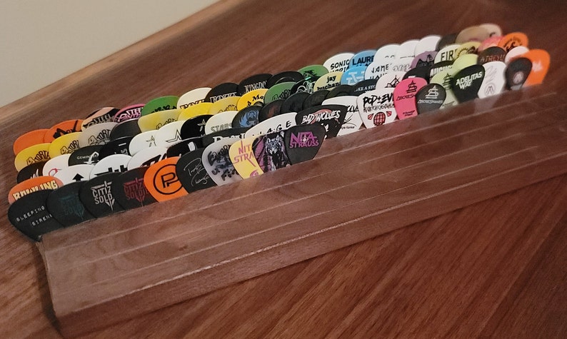 May include: A wooden guitar pick holder with a clear plastic front, displaying a collection of colorful guitar picks with various band logos and names. The picks are arranged in a fan shape, with the holder sitting on a wooden surface.