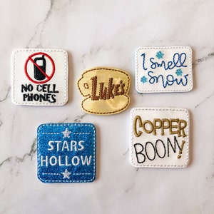 May include: Five embroidered patches with white backgrounds. One patch has a red circle with a diagonal line through a black cell phone and the text "NO CELL PHONES". Another patch has a yellow background with the text "Luke's" in brown. A third patch has the text "I smell snow" in blue with three blue snowflakes. A fourth patch has a blue glitter background with white stars and the text "STARS HOLLOW". The fifth patch has the text "COPPER BOONY!" in gold and black.