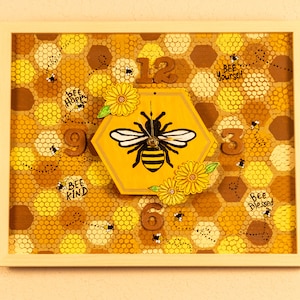 May include: A wooden clock with a honeycomb design and a bee in the center. The clock face has numbers 12, 9, 6, and 3. The clock is surrounded by bees and flowers. The text "Bee Happy", "Bee Yourself", "Bee Kind", and "Bee Blessed" is written around the clock.
