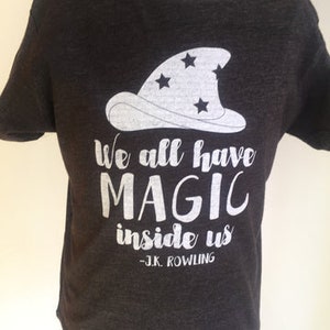 Puede incluir: Una camiseta gris oscuro con un gráfico de sombrero de mago blanco y el texto "We all have magic inside us - J.K. Rowling".