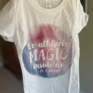 Puede incluir: Camiseta blanca con un diseño de acuarela con la cita "We all have magic inside us - J.K. Rowling".