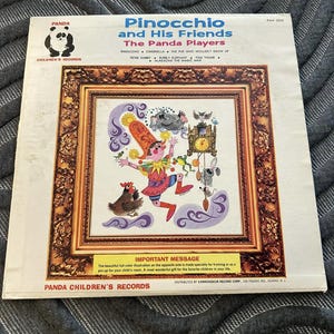 May include: A vintage record album cover featuring Pinocchio and his friends. The cover art is a colorful illustration of Pinocchio with a red hat, a blue shirt, and brown pants. He is surrounded by his friends, including a grey elephant, a white rabbit, a brown chicken, and a grey mouse. The album title is "Pinocchio and His Friends" and the subtitle is "The Panda Players".