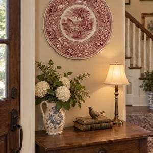 May include: A large round decorative wall plate featuring a detailed red and white transferware landscape scene with a floral border, displayed above a wooden console table with a ceramic pitcher of white hydrangeas and a small bird figurine.