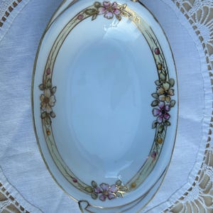 May include: An oval white porcelain serving dish featuring a delicate hand-painted floral border in shades of pink, yellow, and green, accented with fine gold trim along the scalloped edges and a decorative gold-lined rim detail.