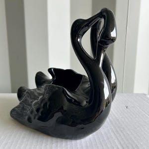 May include: A glossy black ceramic swan figurine featuring a curved neck and a hollowed-out body. The sculpture has a smooth, reflective surface with fluid, molded contours that mimic the shape of a swan in a resting position with wings tucked.