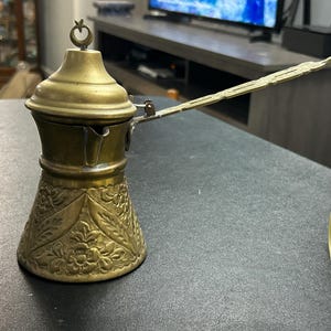 May include: A vintage brass coffee pot with a long handle and ornate floral detailing. The pot has a conical base and a lid with a crescent moon finial. The coffee pot is likely used for making Turkish coffee.