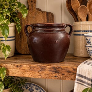 May include: A dark brown, glazed ceramic crock featuring a wide, rounded body and two symmetrical side handles. The vessel has a smooth, reflective surface with a slightly tapered neck and a thick, flat rim, presenting a sturdy, functional design.