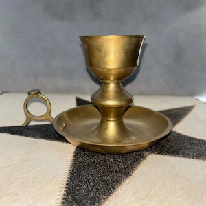 May include: A vintage brass candlestick holder with a circular base and a cup-shaped top. The candlestick has a small handle and is set against a black and white patterned surface. The brass has a warm, golden hue.