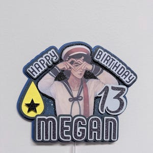 May include: A cake topper with a sailor-themed design. The topper features the text "HAPPY BIRTHDAY 13 MEGAN" and an illustration of a person making a heart shape with their hands. A yellow teardrop shape with a black star is also included.