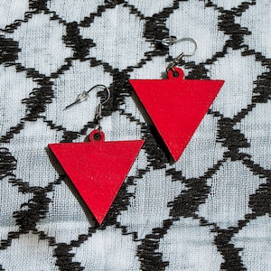 May include: A pair of red triangle shaped earrings with silver hooks. The earrings are on a white and black patterned fabric.