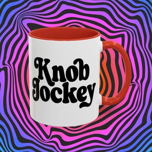May include: White ceramic coffee mug with a red rim and handle. The mug has black text that reads "Knob Jockey" in a retro font.