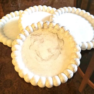 May include: Three white decorative plates with a beaded edge. The plates have a distressed finish with dark gray accents. The plates are round and appear to be made of a ceramic material.