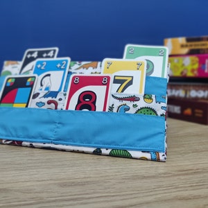 May include: A blue fabric pouch with a white background and colourful dinosaur illustrations. The pouch is filled with various playing cards, including cards with the numbers 2, 7, and 8.