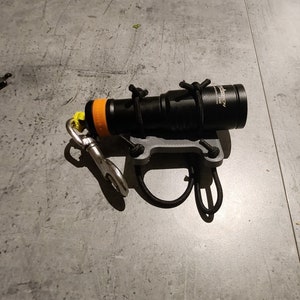May include: Black and orange underwater dive light secured to a gray mount with black rope. A silver clip and yellow cord are attached. The light has the text "Torch" and "Venture" printed on it.