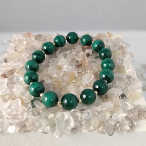 May include: A green malachite beaded bracelet with gold spacers. The bracelet is displayed on a white surface with small clear quartz crystals.