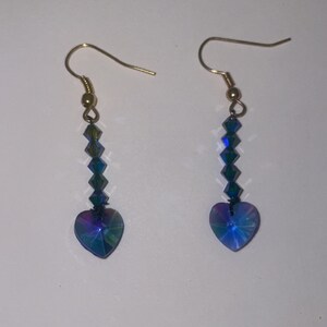 May include: A pair of dangle earrings with a heart-shaped crystal at the bottom. The crystal is iridescent blue and green. The earrings are made with gold-colored metal.