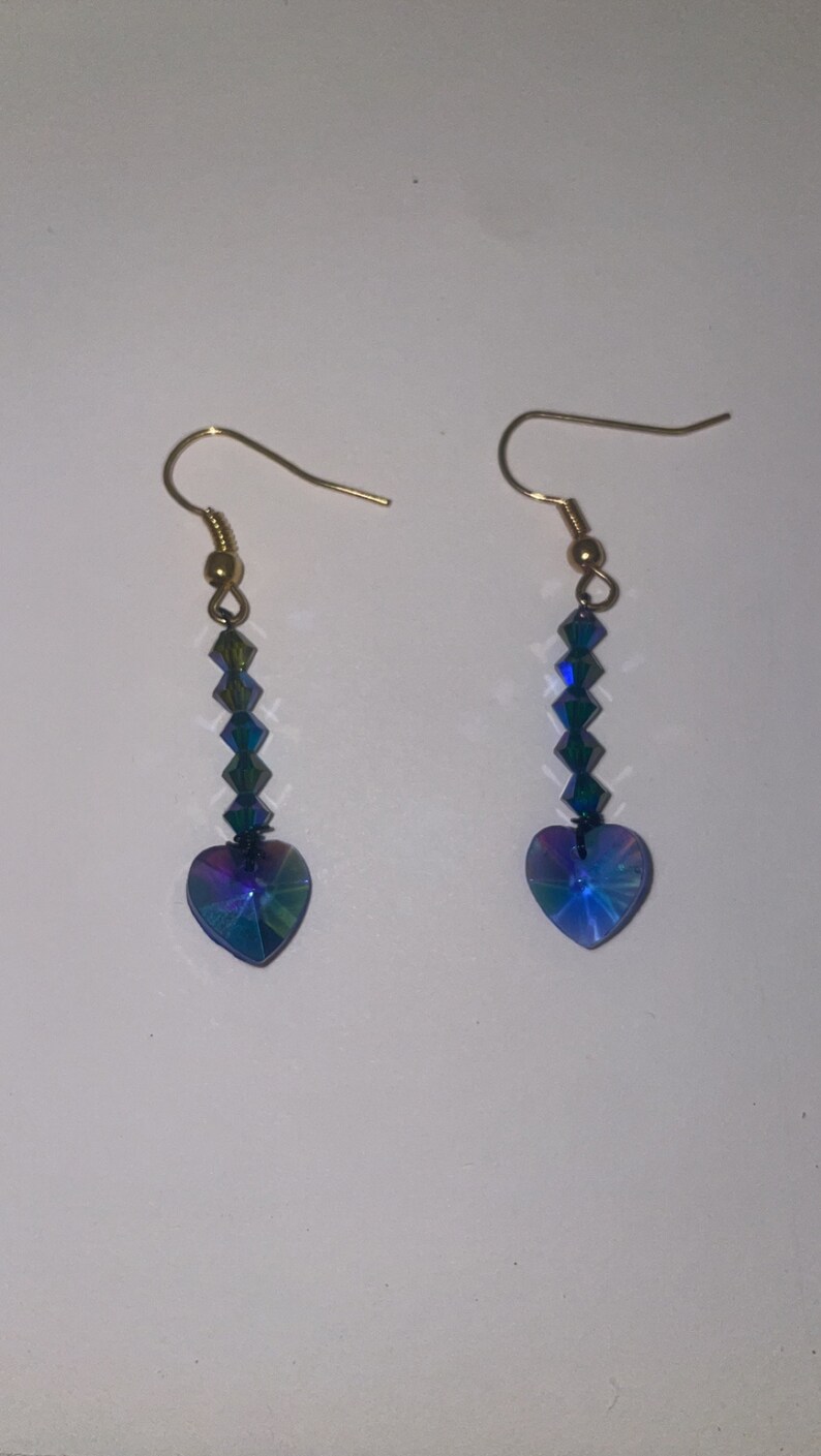 May include: A pair of dangle earrings with a heart-shaped crystal at the bottom. The crystal is iridescent blue and green. The earrings are made with gold-colored metal.