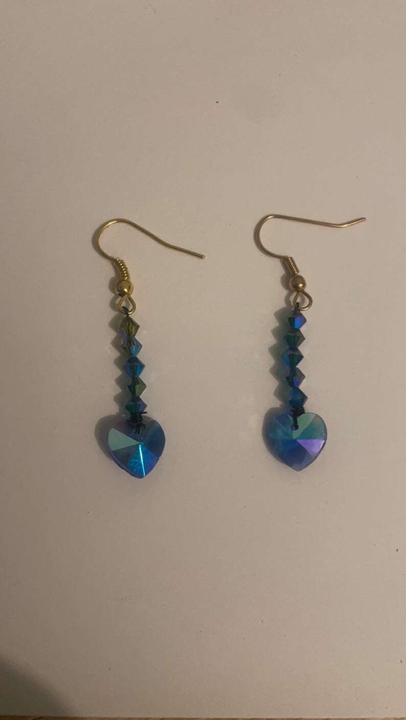 May include: A pair of dangle earrings with a heart-shaped crystal at the bottom. The crystals are iridescent blue and green.