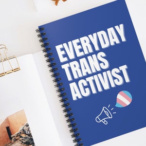 以下が含まれることがあります： 白い文字で「EVERYDAY TRANS ACTIVIST」と書かれた青いスパイラルノート。テキストの下には、青い吹き出しの中に青とピンクのトランスジェンダーのプライドフラッグが入った白いメガホンがあります。