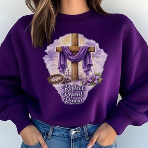 May include: A purple sweatshirt featuring a graphic of a wooden cross draped with purple fabric, a crown of thorns, and flowers. The text "Reflect. Repent. Renew." is below the cross. The sweatshirt is a solid purple color.