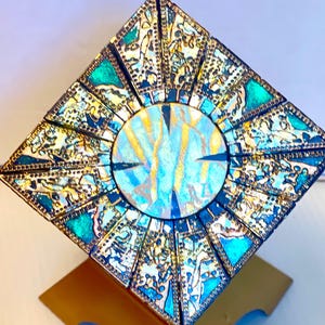 May include: A square, multi-faceted puzzle box featuring a central circular panel with blue, gold, and white swirling patterns. The surrounding triangular panels are decorated with intricate, dark metallic filigree and vibrant teal accents.
