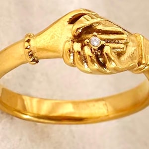 Op de afbeelding: Een goudkleurige ring met twee in elkaar grijpende handen, met een kleine diamant. De ring heeft een gladde, gepolijste band en gedetailleerde handsculpturen. Het ontwerp symboliseert eenheid en partnerschap.