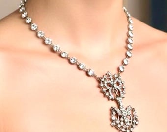 May include: A silver-toned necklace with a sparkling pendant. The necklace features a series of round, clear gemstones. The pendant has an ornate, openwork design with more gemstones. The necklace is worn against a person's skin and a dark blue garment.