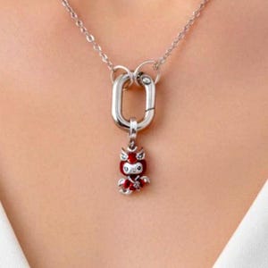 May include: A silver-toned chain necklace featuring a large oval carabiner-style clasp. Attached to the clasp is a small, detailed pendant shaped like a stylised creature with bright red enamel accents and silver-toned metal detailing.