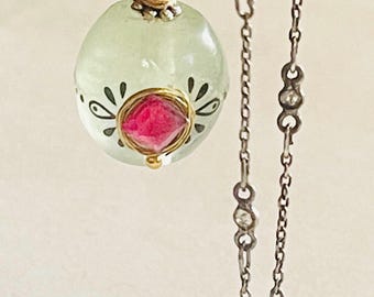 May include: A delicate necklace featuring a light green, round pendant with black floral accents and a pink center stone. The pendant is suspended from a silver chain with additional green and clear stone accents.