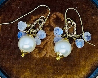 May include: A pair of dangle earrings featuring large white pearl drops with gold accents. The earrings are adorned with small, light blue moonstone beads and gold-toned wire detailing. The earrings are displayed against a dark brown velvet background.