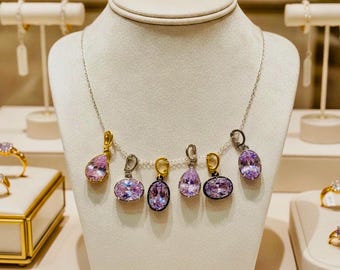 May include: A silver chain necklace featuring six dangling pendants, each set with a faceted light purple gemstone. The pendants vary in shape between oval and pear, with some held in gold-toned settings and others in silver-toned metal settings.