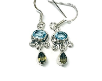 May include: A pair of silver earrings with a blue oval gemstone and a teardrop-shaped green gemstone. The earrings have a decorative swirl design and a hook closure. The background is white.